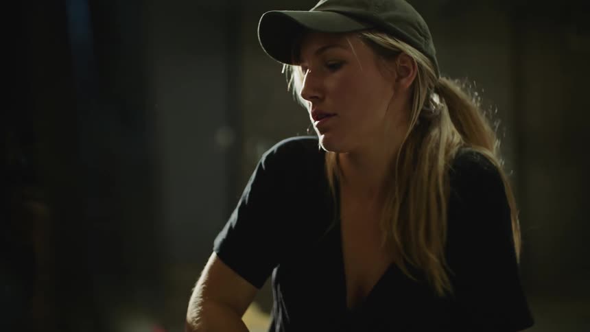 A woman with long blond hair wearing a cap is holding a screwdriver as she works on a motorbike