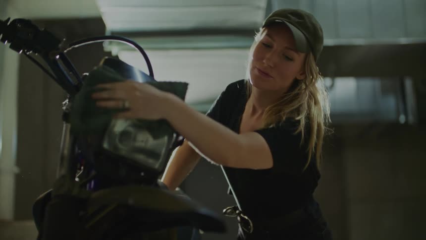 A woman is standing in a garage wiping down a motorcycle with a black cloth