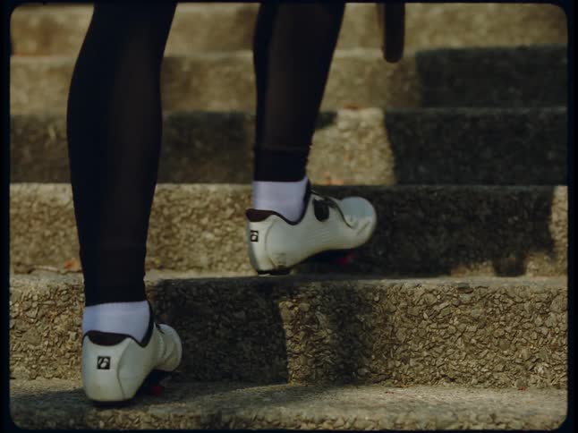 The sun shines on concrete steps as a woman wearing tennis shoes carries a bicycle wheel as she walks up the steps