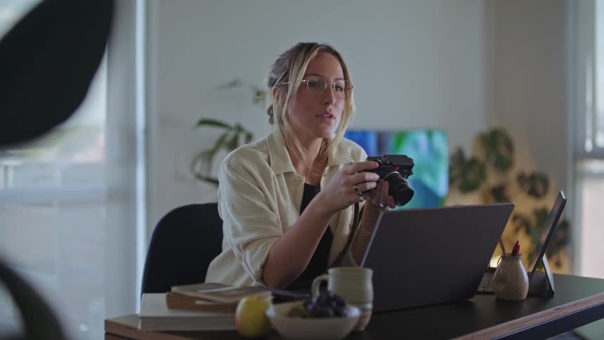 A woman sits at a desk with a laptop in front of her looking at a camera she is holding