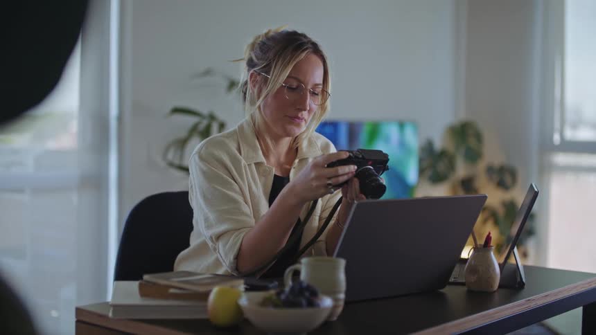A woman is sitting at a table with a laptop computer as she holds her camera and smiles as she looks through pictures