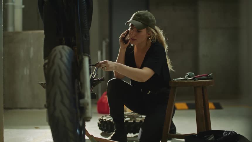 A female mechanic is sitting next to a motocycle she is working on as she holds a screwdriver and talks on her phone