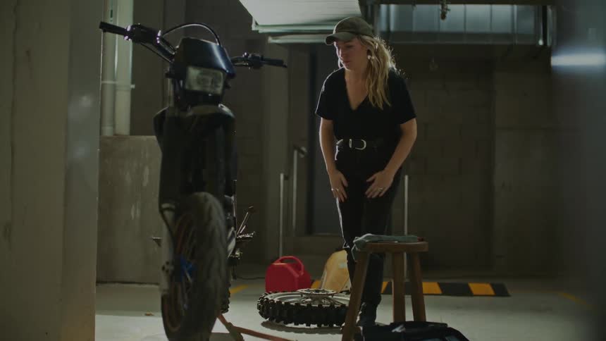 A woman carries a wheel and sets it down near a motorbike as she kneels down and reaches for the frame