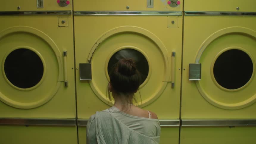 A woman sitting in front of a washing machine facing it as clothes go around in the machine