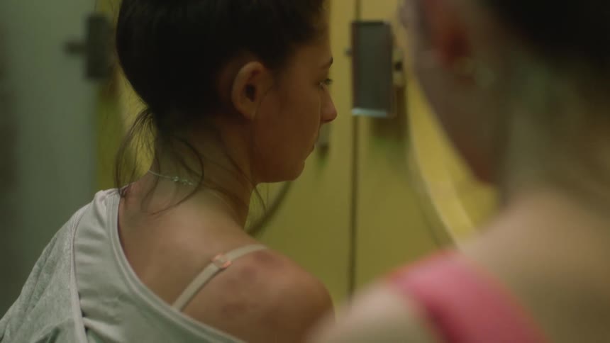 One woman looks toward another who is bruised while they occupy area near machines in laundromat