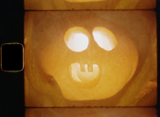 Smoke or haze is coming out of a carved Halloween pumpkin with a wide-open mouth and round eyes and a surprised and shocked facial expression sitting in a tree or bush next to the entry door of a small townhouse on vintage 8mm film