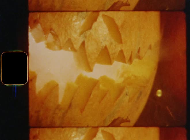 Dynamic 8mm film footage close up shot of the mouth of hollow carved Halloween pumpkin with an evil smiling grimace and aggressive teeth while smoke comes out of the opening that is glowing due to the bright candle in the inside of the jackolantern