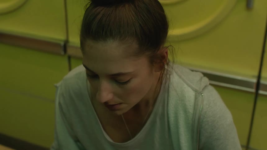 A woman walking in front of washing machines sits down breathing heavily and looking at the floor as another person comes and sits next to her