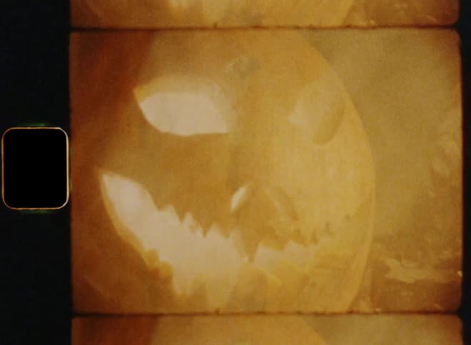Handheld dynamic 8mm film footage revealing a creepy and spooky lit up jack-o-lantern made of a carved pumpkin glowing in the darkness of a fall time night and surrounded by moving smoke causing an unsettling feeling of discomfort and fear