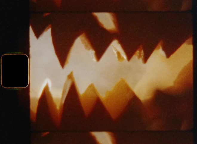 Handheld shaky 8mm film footage closeup of the sharp teeth of a lit up jackolantern made of a carved orange pumpkin with an evil and wicked smile in the darkness at nighttime on Halloween eve