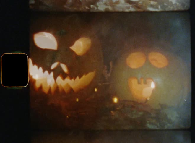 Handheld dynamic 8mm film footage of two Jack O'Lanterns made of carved pumpkins being displayed outdoors for Halloween celebration in flickering flashlight surrounded by smoke from the burning candles or fires to scare kids doing trick or treat