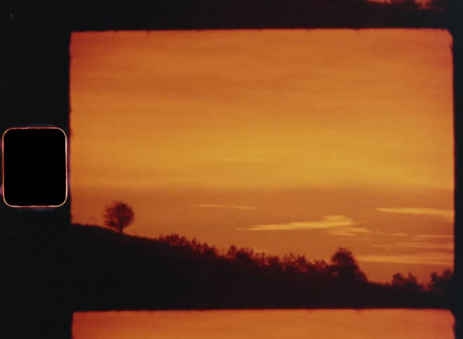 The dark and contrasting silhouette of a hillside with bushes and trees at the horizon in front of an orange evening sky with vibrant afterglow from a stunning sunset and clouds with sepia tones on handheld retro film footage