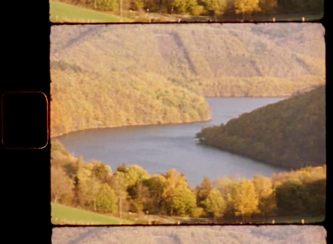 Super 8 mm film footage panning across calm river or reservoir in a valley surrounded by hillside and mountains with grass and trees on a warm and sunny summer day