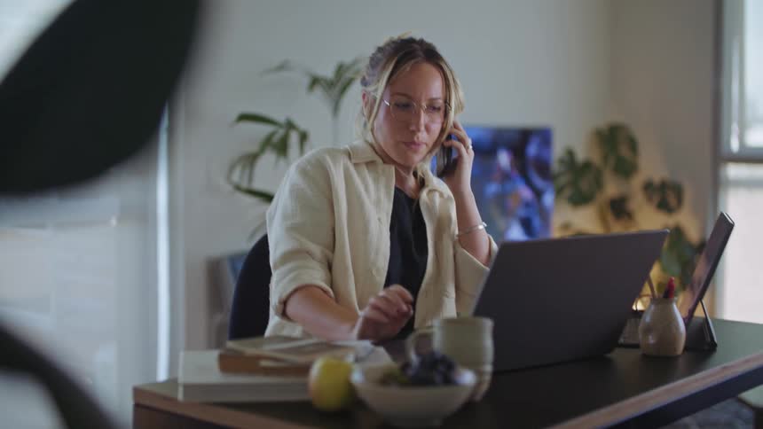 A woman is sitting at her computer desk and talking on the phone