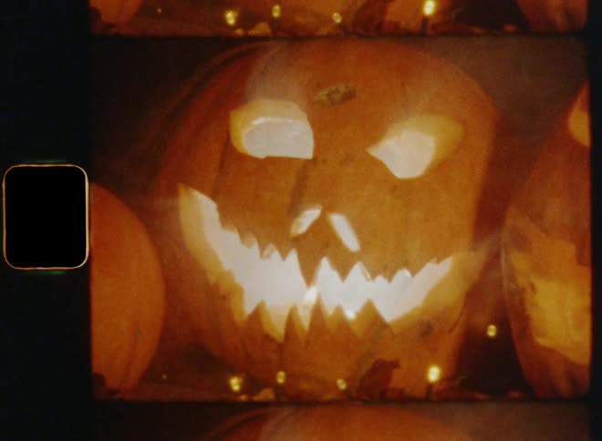 Hand-held shot of three carved pumpkin jack o lanterns standing on a table surrounded by dead oak leaves and a chain of lights as smoke rises from the angry mouths and eyes on nostalgic 8mm film