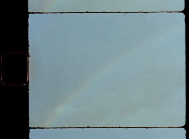 Super 8 mm vintage camera follows a colourful rainbow in front of a bright sky over a landscape with open fields and bushes and grass with handheld movement