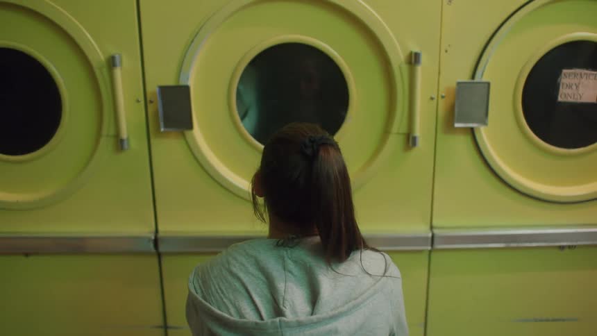 A woman sitting facing a dryer machine as clothes go around in it