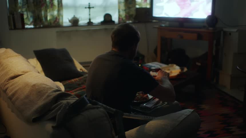 A man sits along on a couch while eating in front of the television