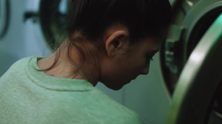 A woman looks apprehensive as she loads clothes into a washing machine at a laundromat