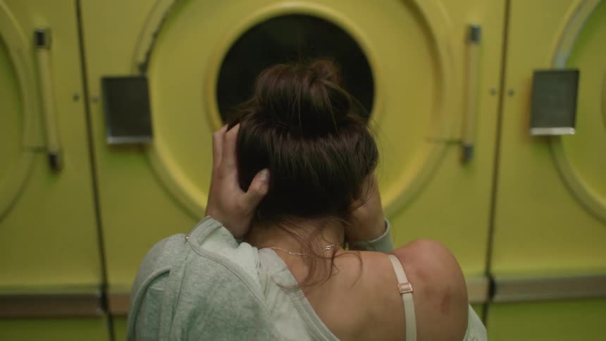 The back side of a young woman standing in front of a dryer at a laundromat hands to head and appears to be crying or screaming