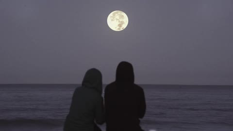 A man and woman sit on the beach under a full moon