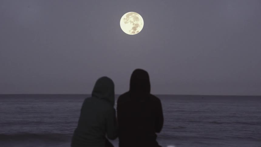 A man and woman sit on the beach under a full moon