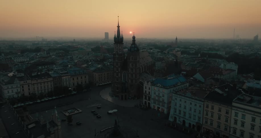 People walk near two towers overlooking a crowded city as tall buildings are spread across town and the sun sets on a foggy horizon
