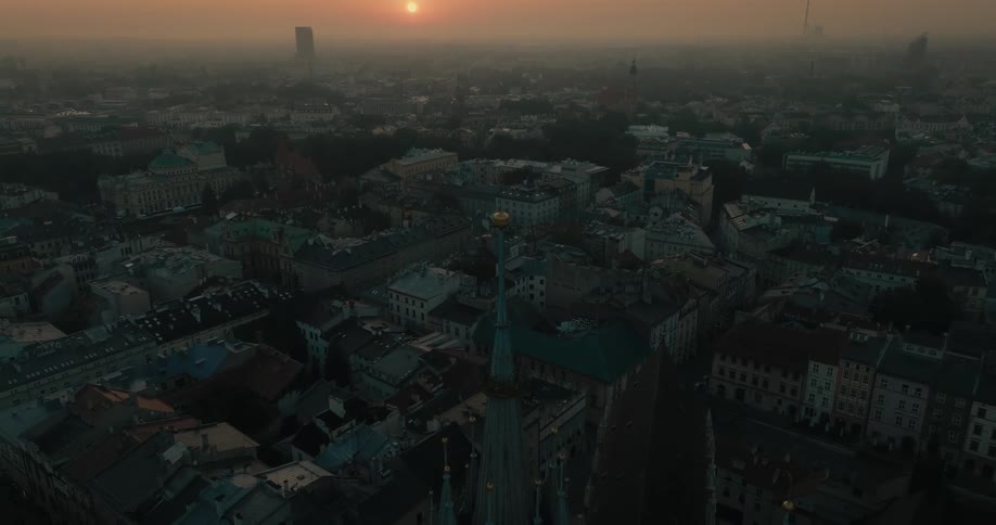 The spire on a tower overlooks a crowded city as tall buildings are spread across town and the sun sets on a foggy horizon