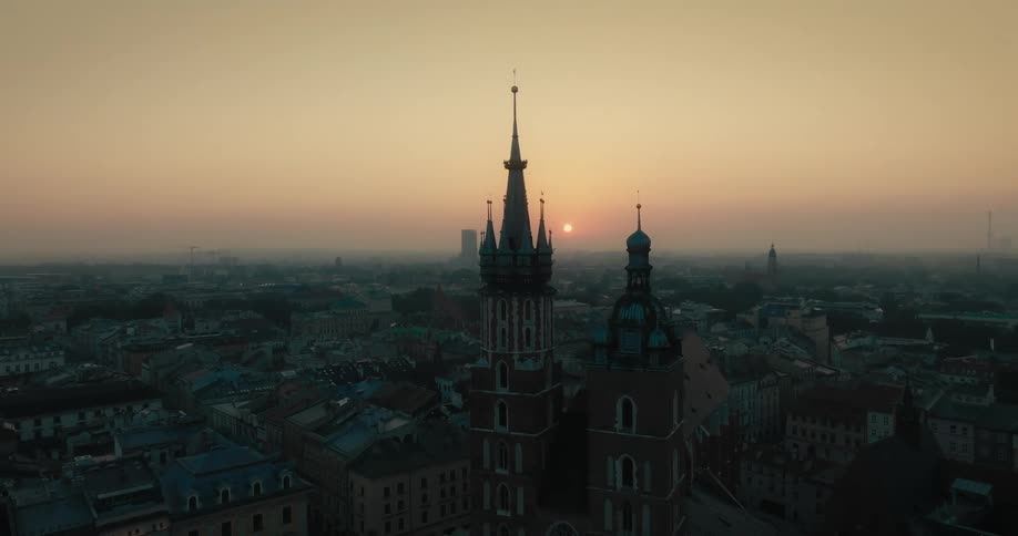 Two towers of a gothic cathedral overlook a crowded city as tall buildings are spread across town and the sun sets on a foggy horizon