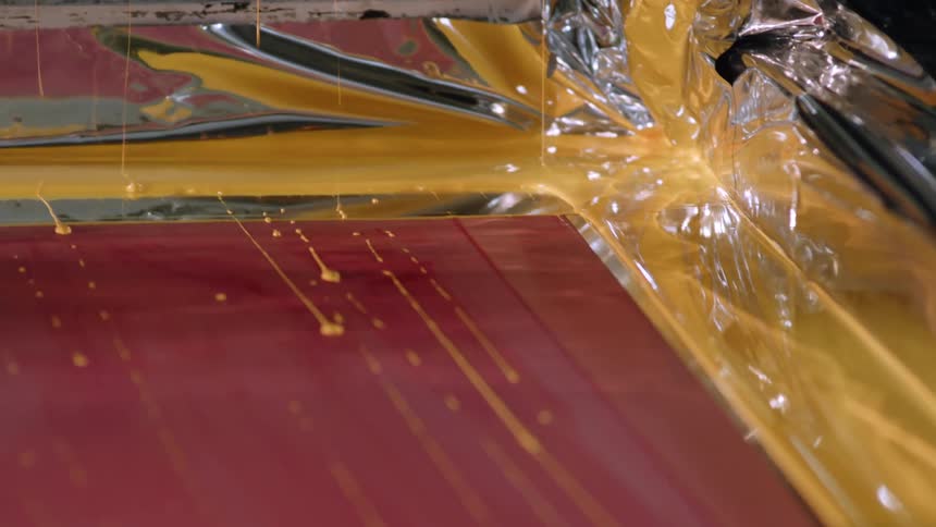 A factory machine moves and yellow paint drips across a red surface