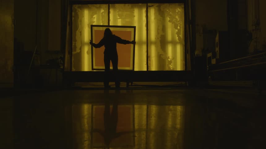 A dark room only lit by yellow window light as a woman carries a frame across the floor and sets it down