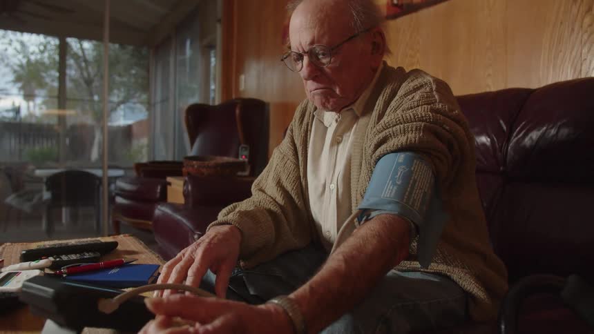 An older man using blood pressure machine at home taps his fingers impatiently and frowns while waiting