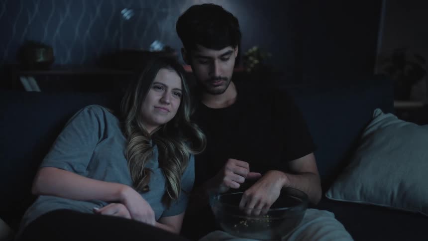 A man and woman sit on a couch leaning against each other while the man finishes a bowl of food and gestures for the woman to get more
