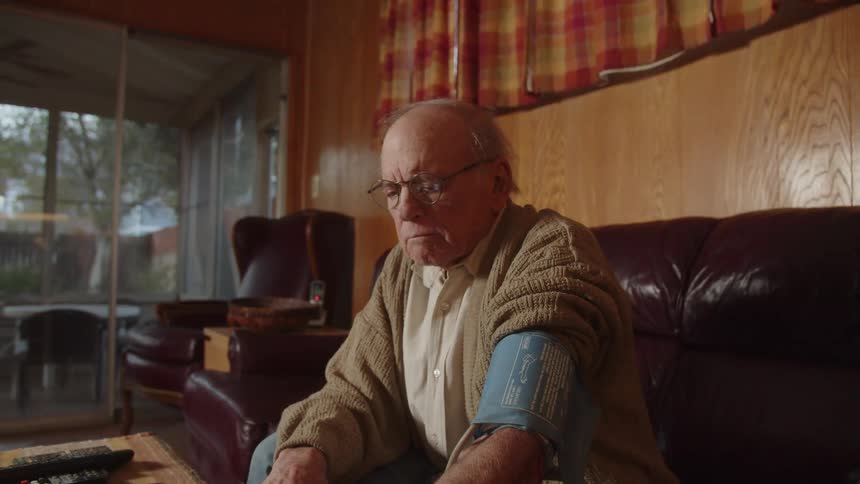 An older man sitting on his couch looking down while he has a blood pressure cuff on his arm