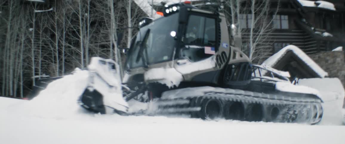 A person driving a snowmobile trail plow through snow in front of a building
