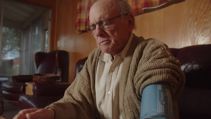 An older man wearing glasses sits on his couch looking sad with a blood pressure cuff on his arm