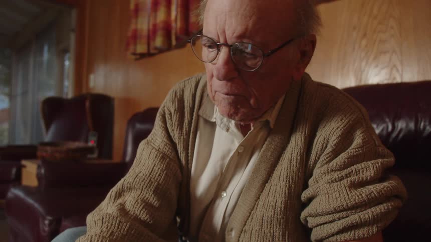 Elderly man with glasses brown jacket and white shirt writes down in notebook sitting on dark brown couch