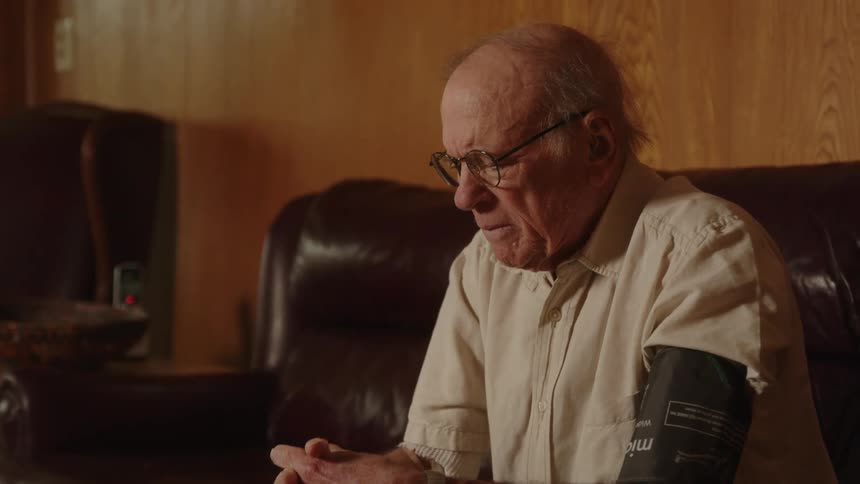 An elderly man sits on a leather chair and looks down at his clasped hands