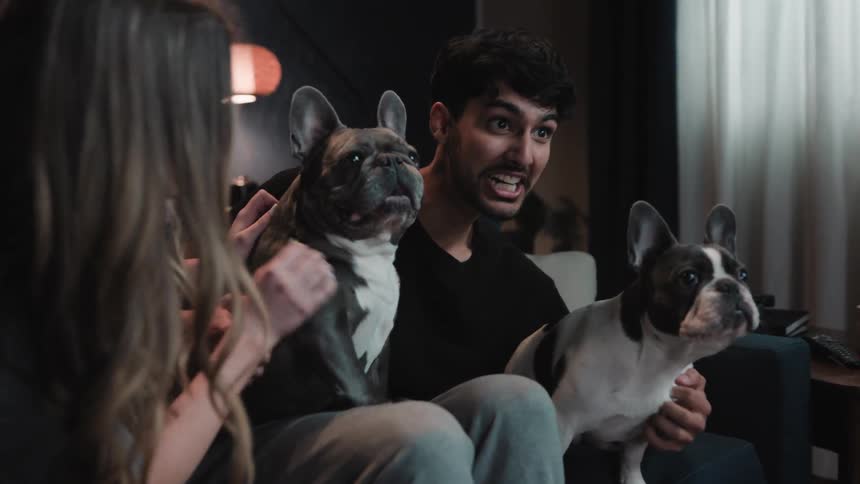 A man sitting on a living room couch is holding two french bulldogs on his lap and making a funny face while the woman next to him is petting one of the dogs