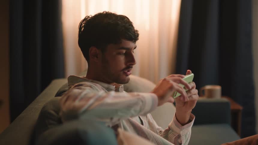 A man is sitting on a couch laughing as he holds his phone and has a conversation