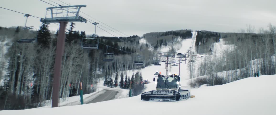 A truck travels up a snow covered hill with ski lifts hanging from cables and other trucks are moving in the distance