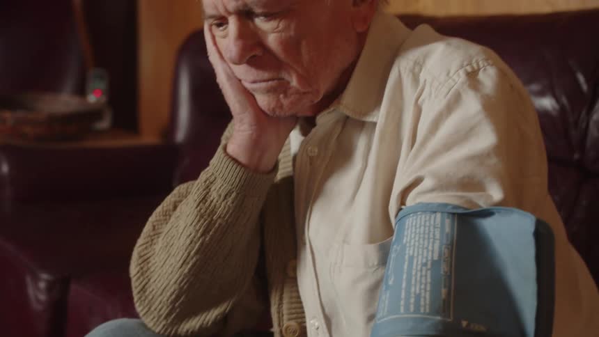 Elderly man with white shirt measures blood pressure sitting on dark brown couch in the living room