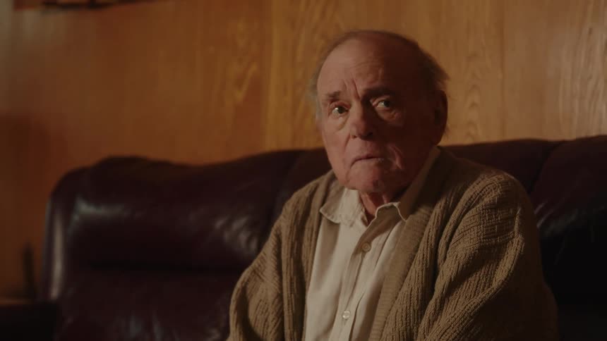 Elderly man with white shirt and brown jacket sits on brown couch and takes off glasses