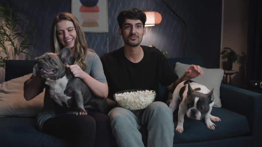 A young man and woman are sitting on a sofa petting their dogs and eating popcorn