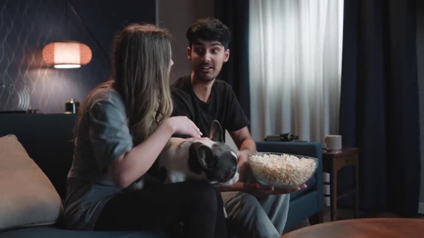 A woman holds a dog on her lap petting it as the man sitting next to her feeds the dog popcorn he is eating