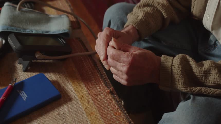 An older man wearing a tan sweater sits on his couch fiddling his hands together
