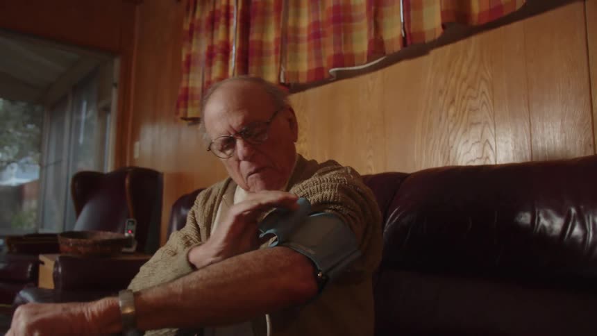 A man is sitting on his living room couch wrapping a blood pressure cuff around his arm