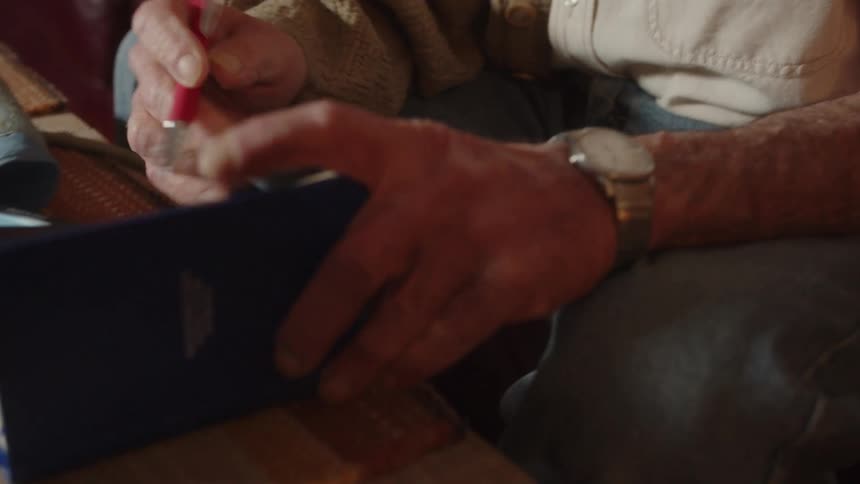 A man is sitting on his living room couch as he opens a notebook near a small machine and he writes on a page