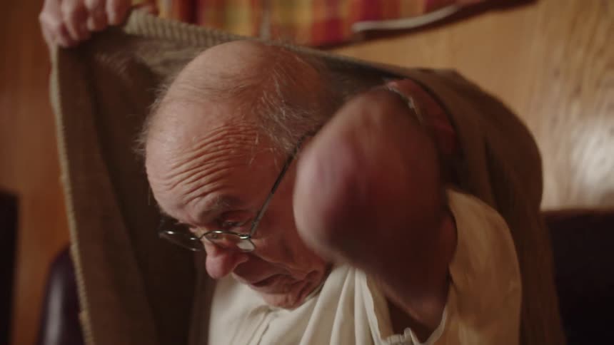 Elderly man sitting on couch puts on sweater before moving items on coffee table around