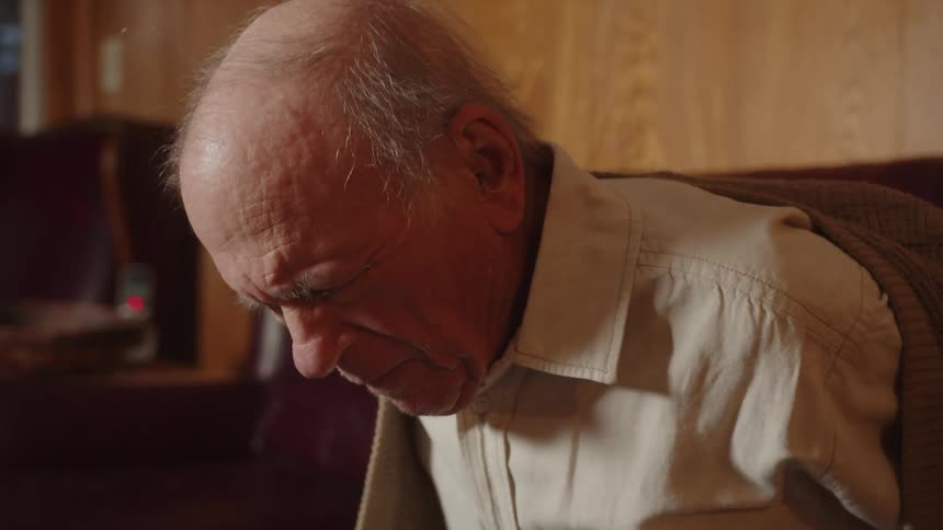 Elderly man sitting on a couch at home takes off a sweater with some difficulty